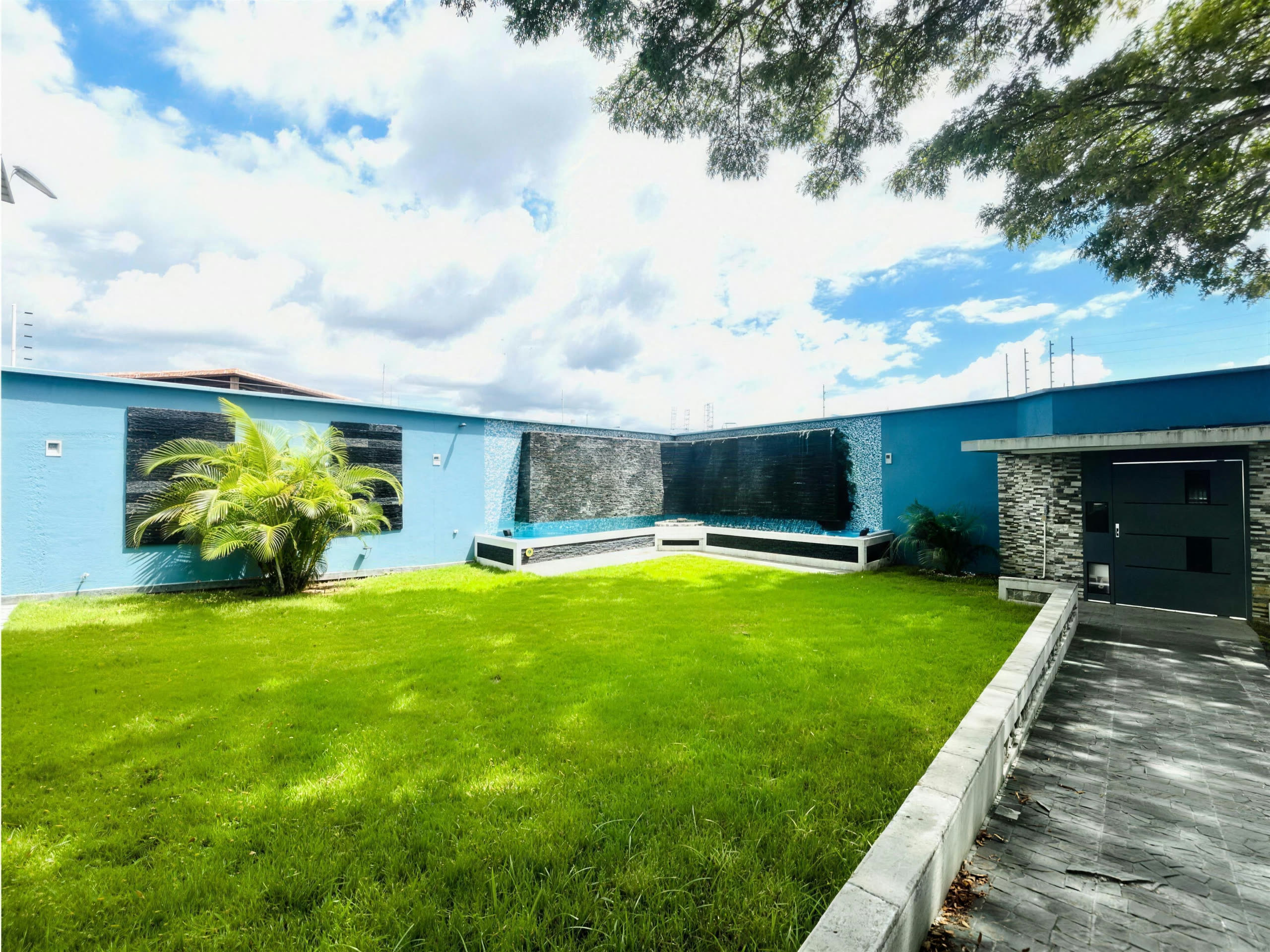 Panorámica del jardín con muro azul, fuente y samán